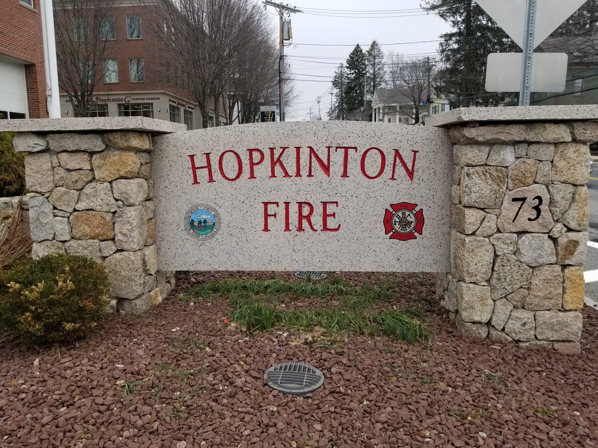 The Hopkinton Fire sign: a speckled granite panel with red-filled lettering, the town seal and a fire service Maltese cross, set between fieldstone piers numbered 73