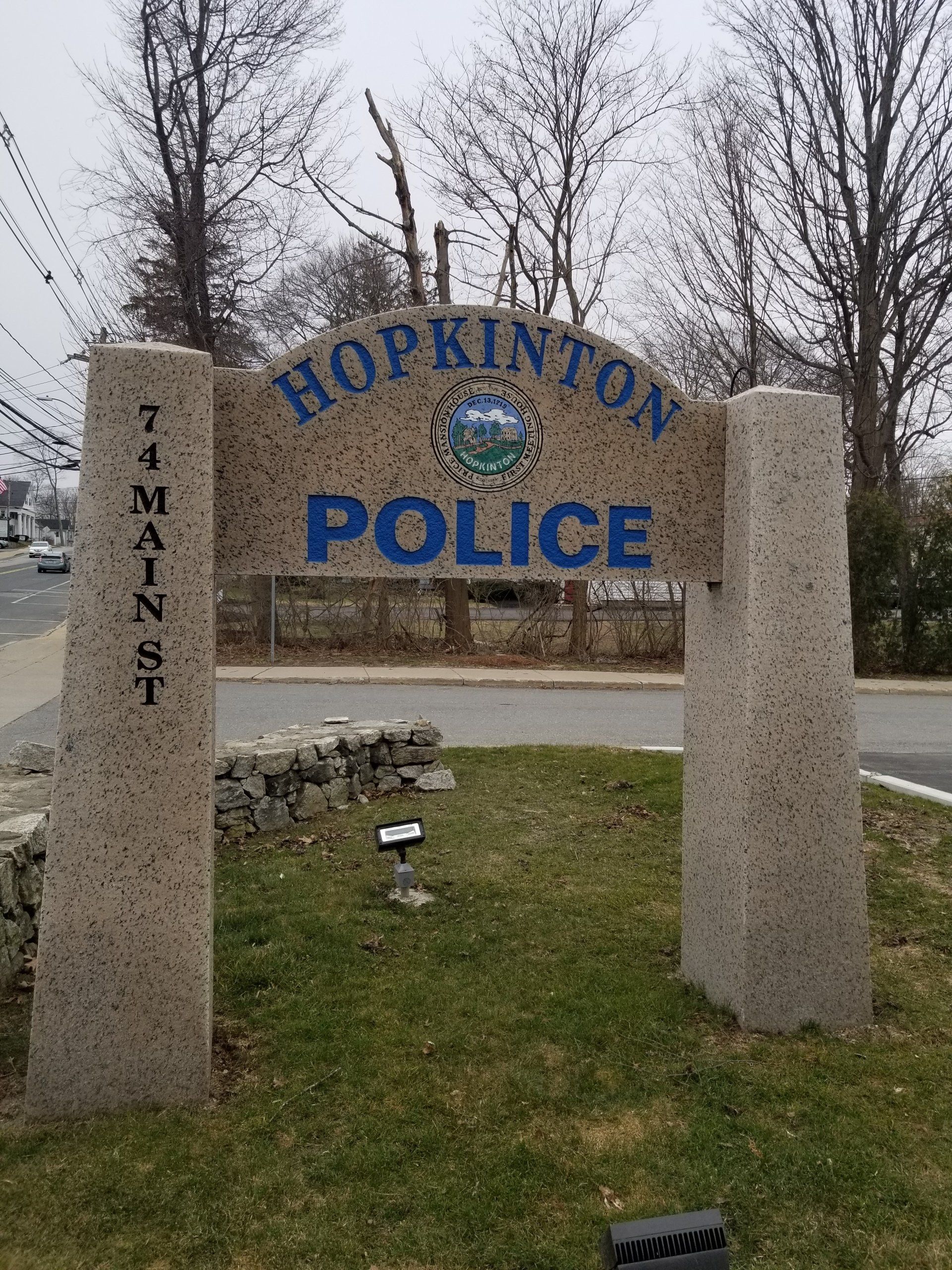 The Hopkinton Police monument: an arched granite crest with blue-filled lettering and the town seal, carried on two tapered granite piers cut 74 Main St