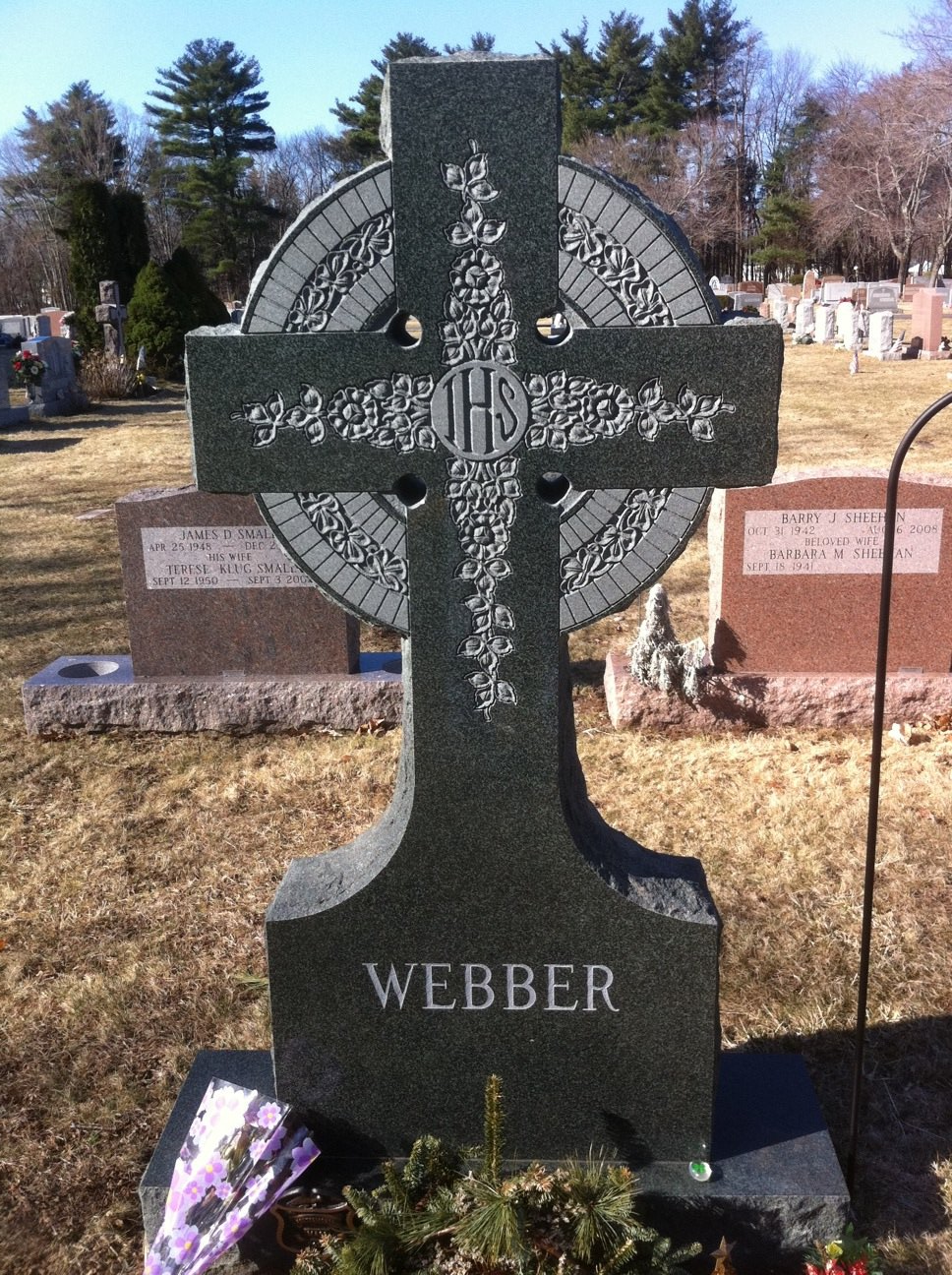 A standing Celtic cross in green granite with a carved ring, floral tracery and an IHS monogram, lettered Webber, standing in a cemetery with pink and grey stones behind it