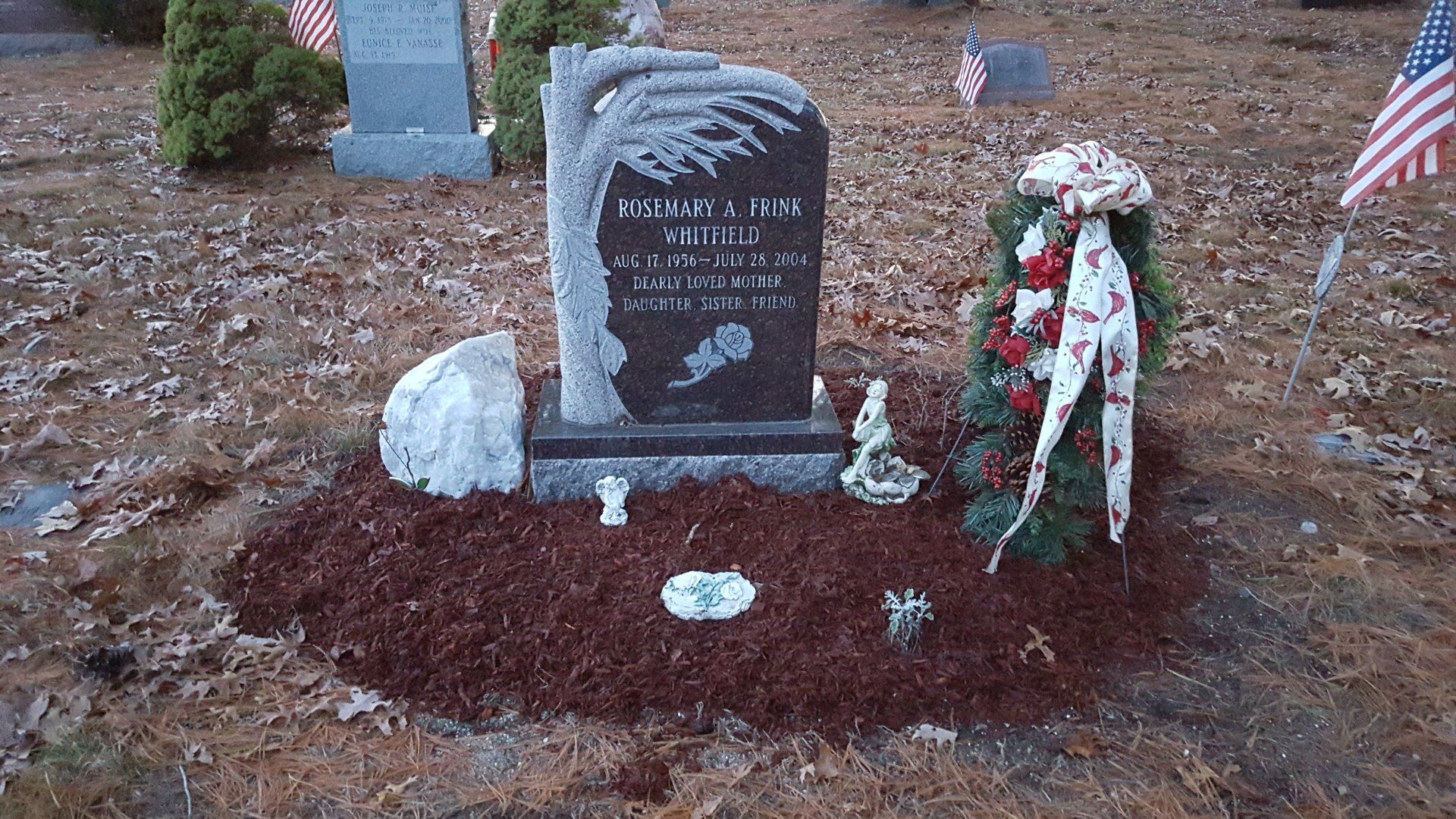 Dark granite upright monument with a tree trunk and branch carved down one side, set in a bed of mulch beside a boulder