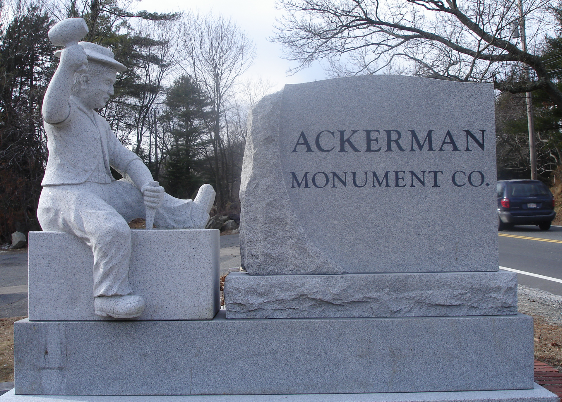 The Ackerman Monument Co. roadside sign: a rough granite boulder lettered ACKERMAN MONUMENT CO. beside a life-size granite carving of a stonecutter in a cap raising his mallet, chisel in hand